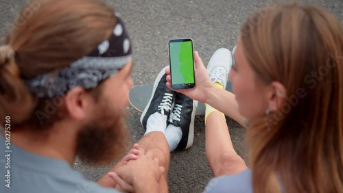 Beauty Young millennial friends sitting outside and looking at green screen phone in park. Happy smiling people using modern smartphone technology. Media lifestyle, internet chat video live conferance photo