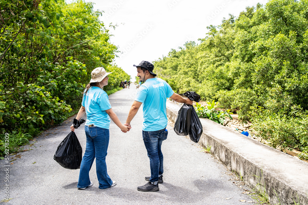 Asian couple walks hand in hand collecting rubbish in a forest path, Young man and woman wearing uniforms, gathering glass bottles into plastic bags. Nature care concept Asian couple walks hand in hand collecting rubbish in a forest path, Young man and woman wearing uniforms, gathering glass bottles into plastic bags. Nature care concept