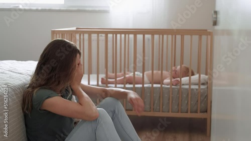 Young mother suffering postnatal depression sitting on floor next to baby napping in bed, resting head on knees, crying, feeling desperate in need of professional psychological support photo