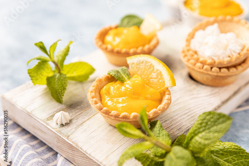 Shortbread tartlet filled with lemon curd, mint and lemon slices and mini meringue on wooden board, blue background.