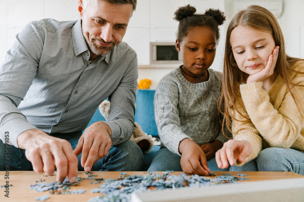 Fototapeta premium Adopted african little girl solving puzzles with father and sister