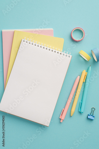 School supplies concept. Top view vertical photo of colorful stationery stack of notebooks pens binder clip and adhesive tape on isolated pastel blue background with blank space