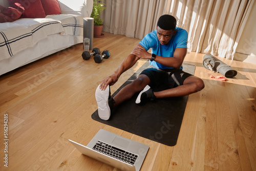 Young black African American young male working out at home stretching his legs before following an online exercise class in his living room on his laptop
