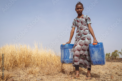 Fotografiet African teenager with two blue water cans walking through a field of dried up gr