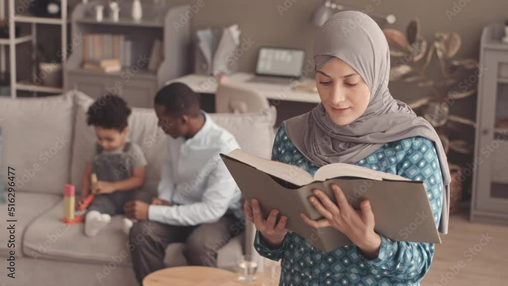 Medium slowmo portrait of young Muslim woman in grey hijab reading Quran at home while her African American husband playing with their little son on sofa in background Medium slowmo portrait of young Muslim woman in grey hijab reading Quran at home while her African American husband playing with their little son on sofa in background