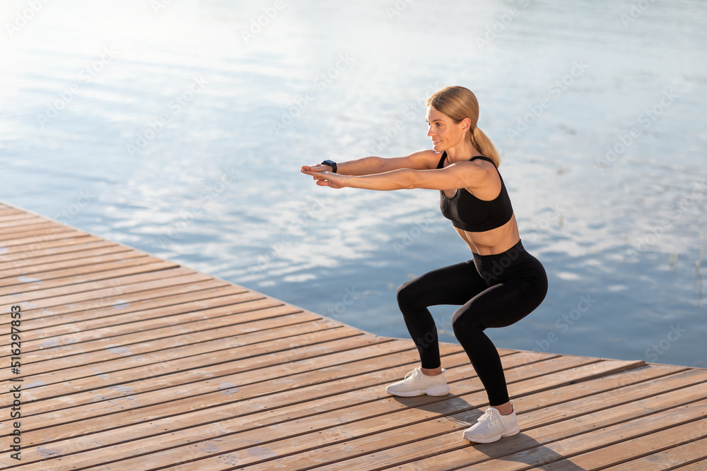 Outdoor Training. Smiling middle aged woman making squats exercise on the pier,