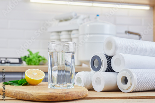 Glass of filtered clean water with reverse osmosis filter, lemons and cartridges on table in kitchen. Concept Household filtration or purification system.