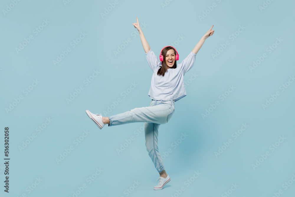 Full body young happy fun woman she 20s wear casual blouse headphones listen to music dance raise up hands leg isolated on pastel plain light blue background studio portrait. People lifestyle concept