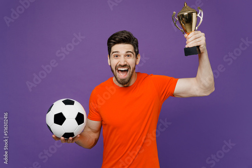 Young exultant overjoyed excited fan man he wears orange t-shirt cheer up support football sport team hold in hand soccer ball champion cup watch tv live stream isolated on plain purple background.