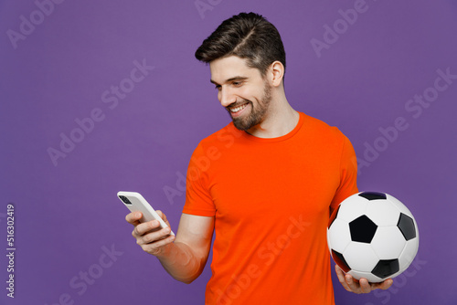 Young fun fan smiling happy man he wears orange t-shirt cheer up support football sport team hold in hand soccer ball watch tv live stream use mobile cell phone isolated on plain purple background