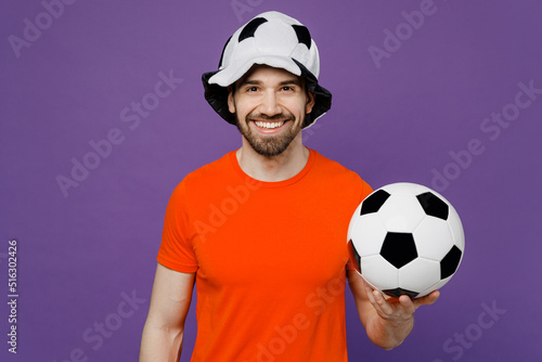 Young smiling cheerful happy cool fun fan man he wears orange t-shirt cheer up support football sport team hold in hand soccer ball panama hat watch tv live stream isolated on plain purple background. © ViDi Studio