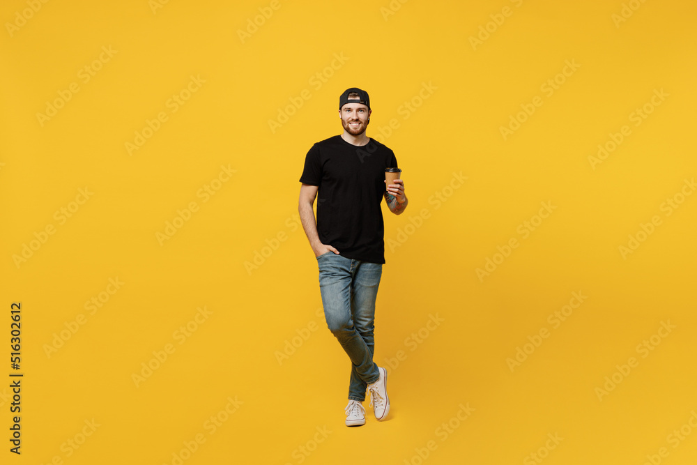 Full body young bearded tattooed man 20s he wears casual black t-shirt cap hold takeaway delivery craft paper brown cup coffee to go isolated on plain yellow wall background. People lifestyle concept. Full body young bearded tattooed man 20s he wears casual black t-shirt cap hold takeaway delivery craft paper brown cup coffee to go isolated on plain yellow wall background. People lifestyle concept.