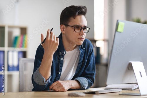 Confused male office employee sitting at wokplace and looking at computer screen photo