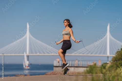 Girl jumps on a rope in a park against the background of a bridge across the bay. Morning workout of a Spanish girl on the beach. A young European woman does sports in nature on the weekend. Health. © Iona