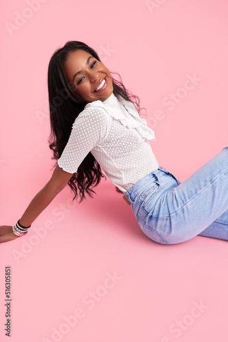 Trendy young ethnic female model smiling while sitting in pink studio