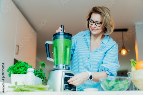 Middle aged woman with blender and green vegetables making detox shake or green smoothie at home. Healthy dieting, eating, cooking. Natural anti aging, weight loss program. Vegan, vegetarian diet.