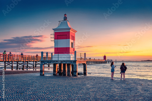 Fototapeta Naklejka Na Ścianę i Meble -  Tourists on the shore of Lignano Sabbiadoro resort. Splendid outdoor scene of Adriatic coast of Italy with lighthous and old wooden pier. Picturesque morning seascape of Mediterranean sea.