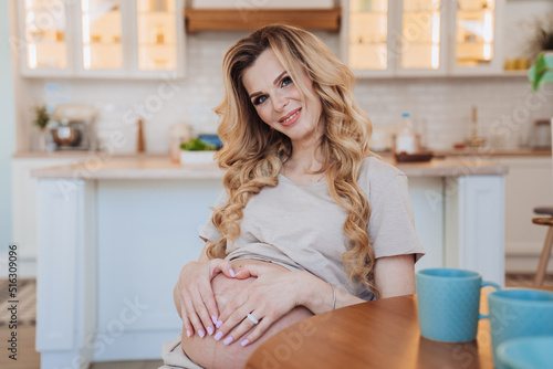 Adorable pregnant blonde swedish woman sitting at table at kitchen forming heart by hands on her belly, happy smiling waiting for a new life. Beautiful caucasian pregnant housewife at home. Happy life