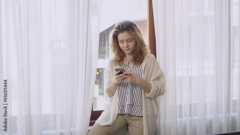 young woman enjoy using mobile, talk with friend on mobile phone. lady sitting alone near window curtain at home in holiday young woman enjoy using mobile, talk with friend on mobile phone. lady sitting alone near window curtain at home in holiday
