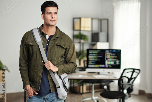 Portrait of young ambitious software developer with cross body bag standing in office and looking away photo