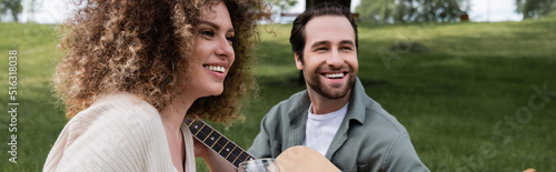 romantic man playing acoustic guitar near curly woman during picnic, banner. © LIGHTFIELD STUDIOS