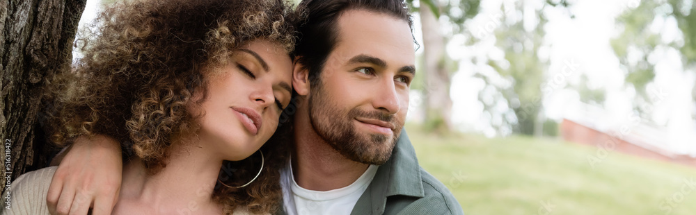 happy man embracing curly and young woman with closed eyes near tree trunk, banner. happy man embracing curly and young woman with closed eyes near tree trunk, banner.