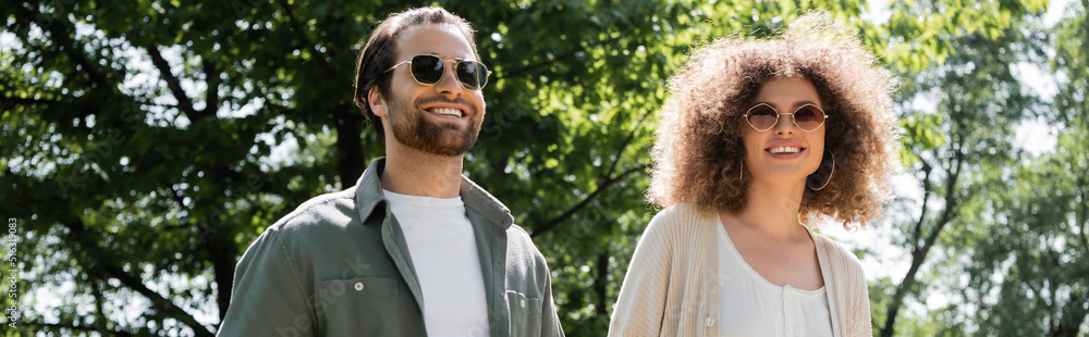 curly woman and happy man in stylish sunglasses walking in park, banner. curly woman and happy man in stylish sunglasses walking in park, banner.