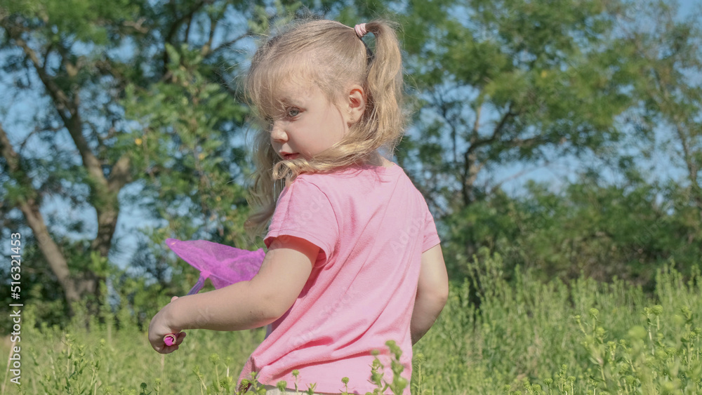 Little girl plays with butterfly net of tall grass in city park. Cute little girl is playing with aerial insect net in meadow on sun day. Little girl plays with butterfly net of tall grass in city park. Cute little girl is playing with aerial insect net in meadow on sun day.