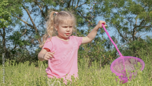 Little girl plays with butterfly net of tall grass in city park. Cute little girl is playing with aerial insect net in meadow on sun day.