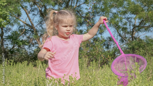 Little girl plays with butterfly net of tall grass in city park. Cute little girl is playing with aerial insect net in meadow on sun day.