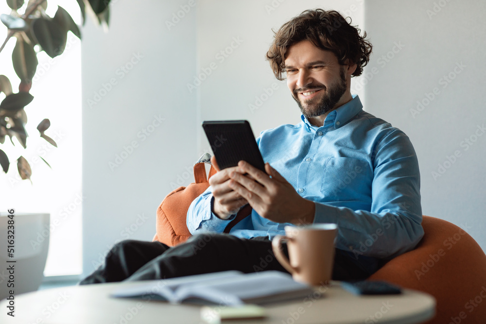 Happy mature businessman using tablet pc, looking at screen, sitting on bean bag chair in modern office, empty space
