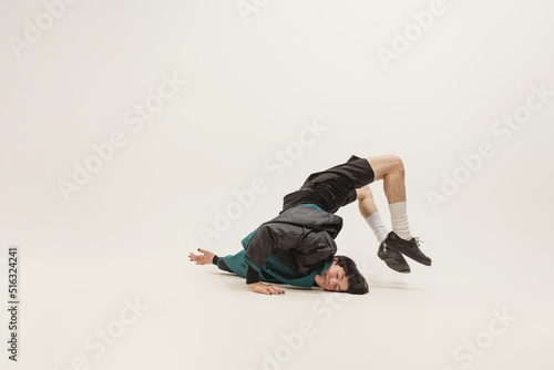 Stylish young man in black outfit and green coat posing, lying on floor isolated over grey studio background. Flexibility.