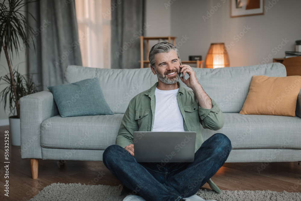 Smiling caucasian retired male with beard sits on floor with laptop, calls by phone in living room Smiling caucasian retired male with beard sits on floor with laptop, calls by phone in living room