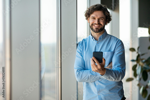 Happy mature enterpreneur browsing internet on smartphone, standing near big window in modern office, copy space photo