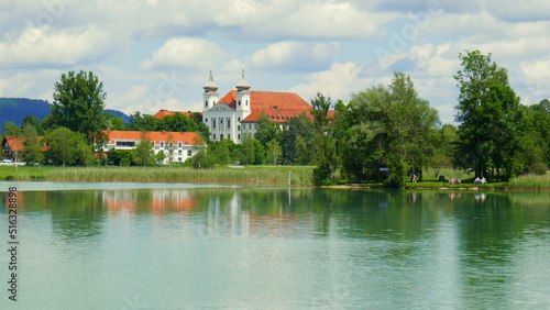 malerischer Blick auf Ufer des Kochelsees mit Kirche von Schlehdorf, Schilf, Wald, Menschen und Wolken