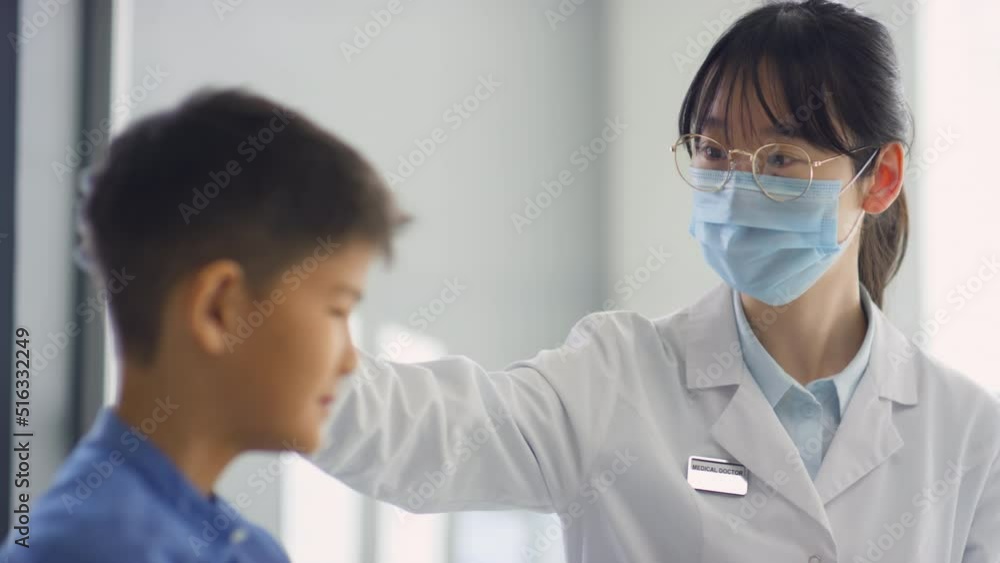 Asian female doctor in protective mask and gloves stroking head of sick kid while supporting him in hospital Asian female doctor in protective mask and gloves stroking head of sick kid while supporting him in hospital