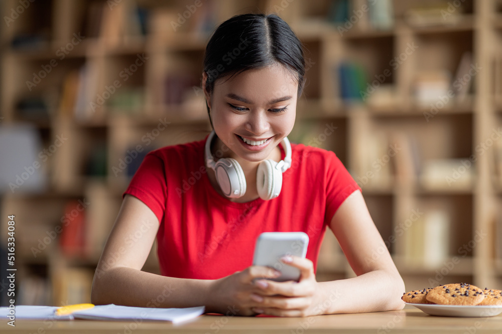 Glad smiling adolescent japanese girl in headphones typing on smartphone, have social media chat Glad smiling adolescent japanese girl in headphones typing on smartphone, have social media chat