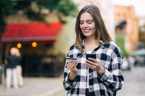 Cheerful young beautiful woman using smartphone and credit card to pay on the street.