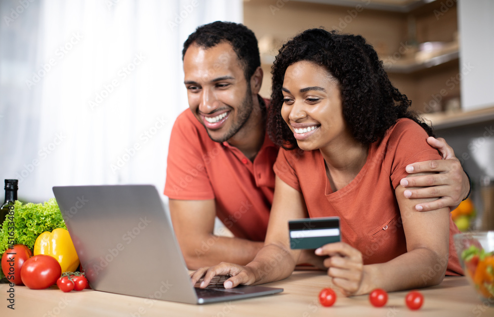 Cheerful young african american wife and husband at table with laptop, credit card and organic vegetables Cheerful young african american wife and husband at table with laptop, credit card and organic vegetables