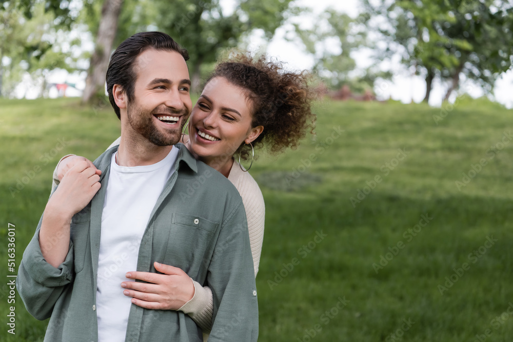 pleased and curly woman hugging boyfriend from behind in green summer park. pleased and curly woman hugging boyfriend from behind in green summer park.