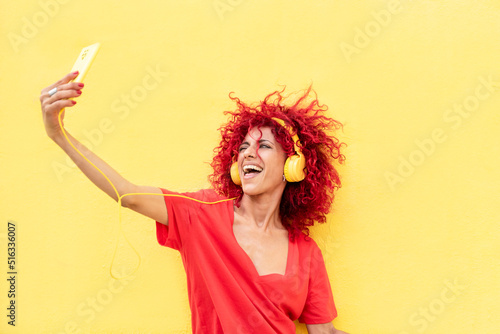 a beautiful smiling latin woman with red afro hair listen to music with yellow headphones and taking a selfie with a yellow smartphone on a yellow background