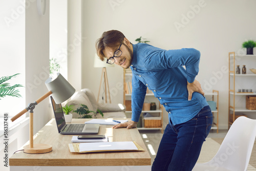 Male office worker experiences severe back pain caused by prolonged sitting in wrong position. Unhappy man experiencing spasm of spine getting up from behind workplace in office.Sedentary life concept