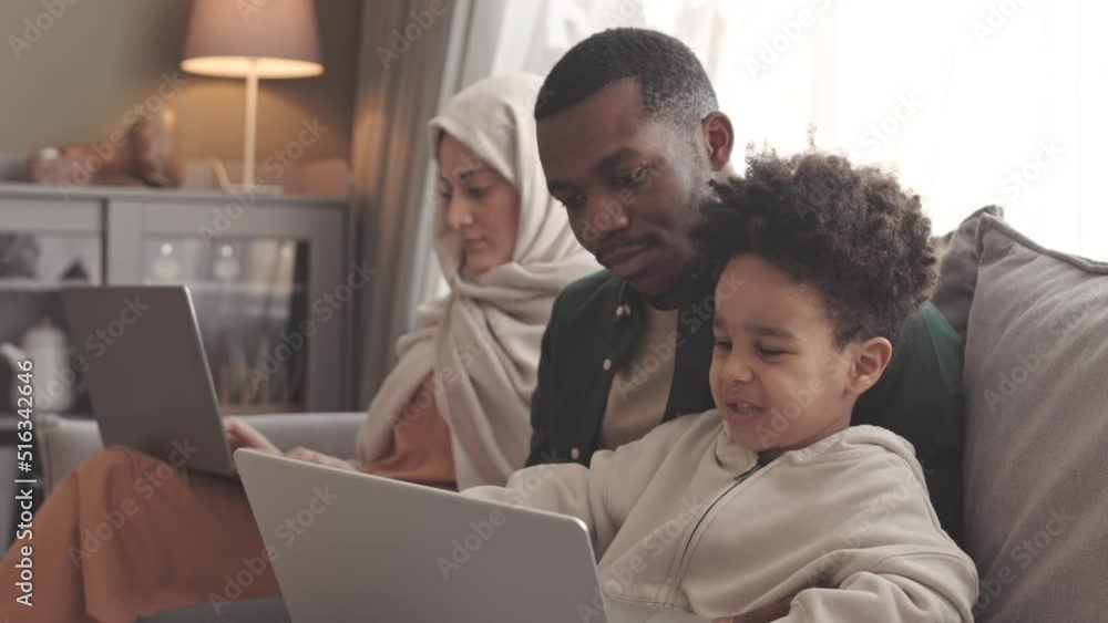 Slowmo of African American Muslim father and his toddler son watching cartoons on laptop while his wife in headscarf working on laptop sitting on sofa nearby Slowmo of African American Muslim father and his toddler son watching cartoons on laptop while his wife in headscarf working on laptop sitting on sofa nearby