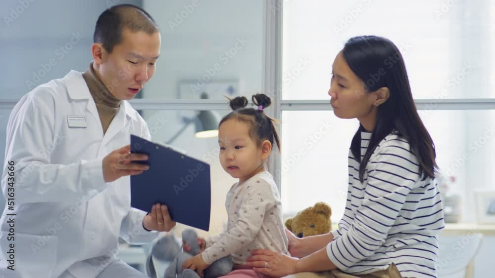 Asian pediatrician showing papers on clipboard to playful toddler girl and speaking with her mother during consultation in clinic Asian pediatrician showing papers on clipboard to playful toddler girl and speaking with her mother during consultation in clinic