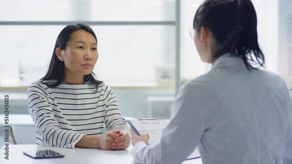 Asian woman giving medical history to female doctor and talking with her during consultation in clinic Asian woman giving medical history to female doctor and talking with her during consultation in clinic