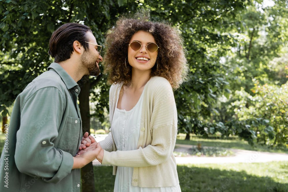 cheerful woman holding hands with happy man in stylish sunglasses in park. cheerful woman holding hands with happy man in stylish sunglasses in park.