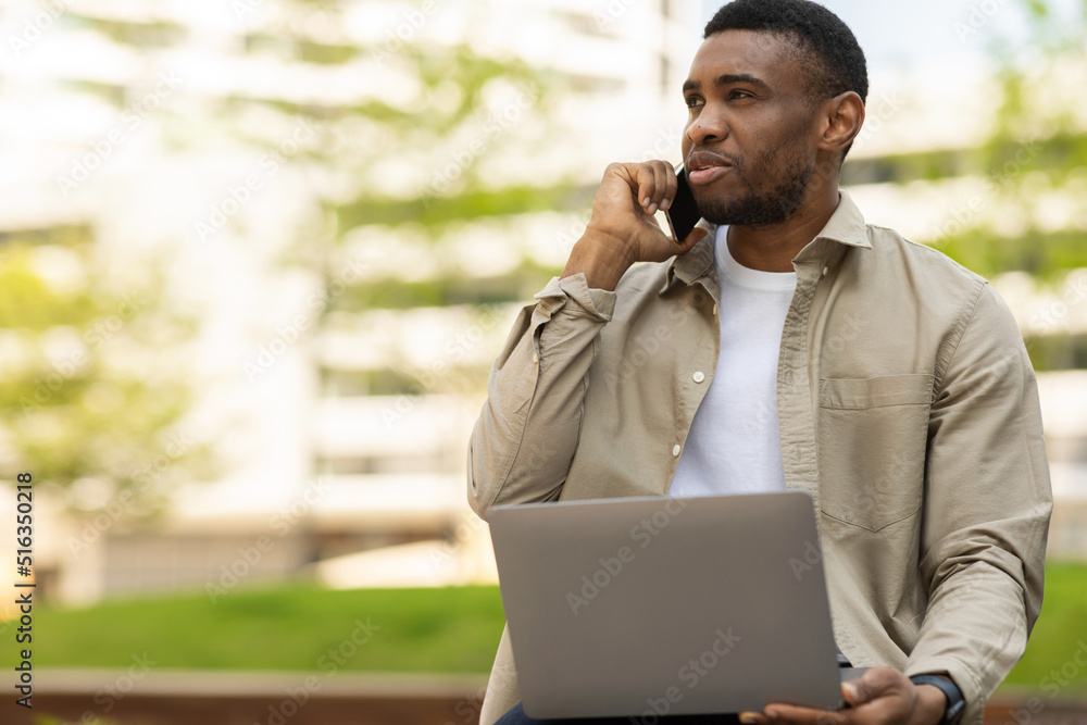 A black man is talking on a mobile phone and working on a laptop on a bench against the backdrop of the city A black man is talking on a mobile phone and working on a laptop on a bench against the backdrop of the city