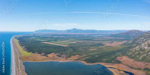 Aerial view of the sea coast and valley among the mountains. Agricultural fields in the distance. In the foreground is the shore of the lake. Nature of Siberia. Olsky district, Magadan region, Russia.