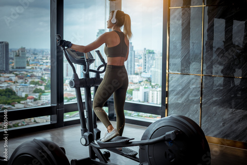 Young fitness sport woman using an elliptic machine trainer at gym fitness center, listening a music with headphones, machine aerobic for slim and firm healthy lifestyle.