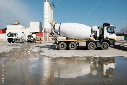 White concrete mixers standing by a modern concrete plant. Reflections of vehicles in puddle of water.